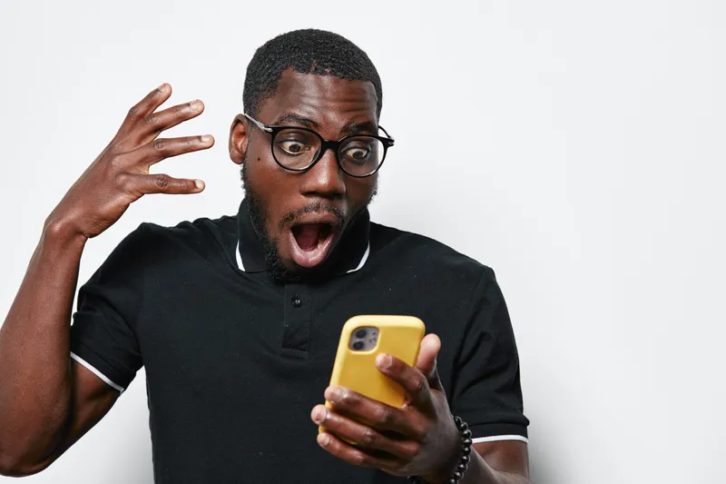 African American man with dark skin and glasses looking shocked at yellow smartphone in black polo shirt on white background. Surprised expression and casual style.