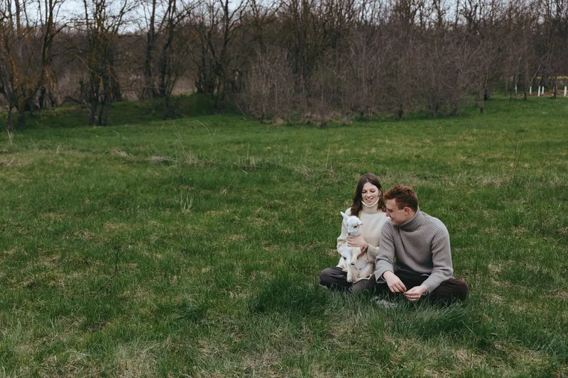 Young couple sits on green grass in a quiet park holding a small white baby goat enjoying a peaceful spring day together with casual warm clothing and relaxed expressions