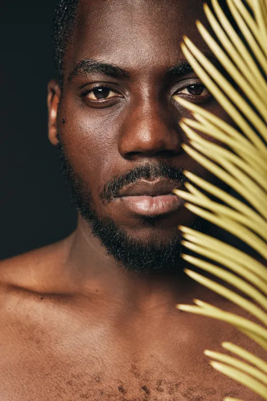 African man with dark skin and beard in creative conceptual visual style, closeup portrait with palm leaf, natural texture and artistic expression for beauty and identity.