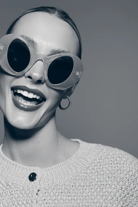 Happy Caucasian woman with light skin wearing large round sunglasses and earrings smiles broadly in studio. Close-up portrait with joyful expression and cozy sweater.