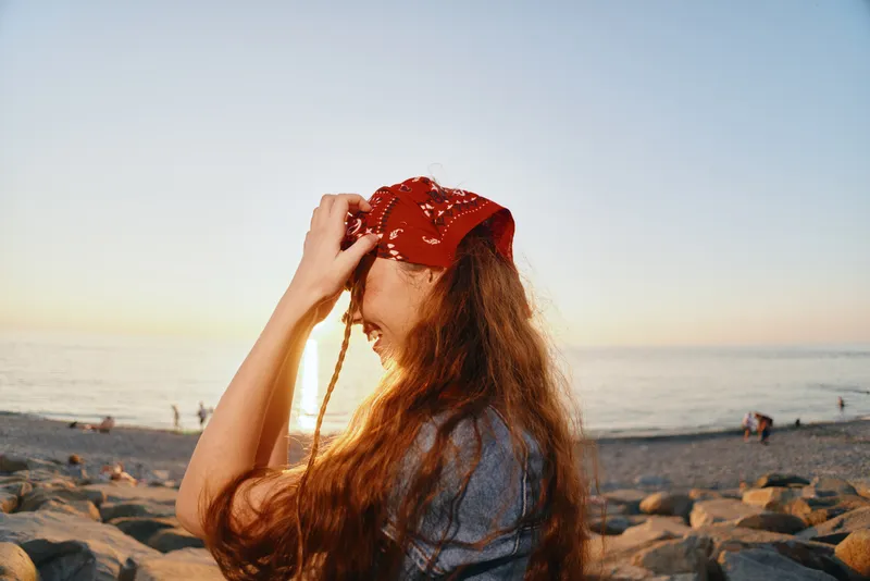 Lifestyle woman in modern boho-western denim street style wearing a red bandana, smiling by the beach during golden hour with warm film color tones.