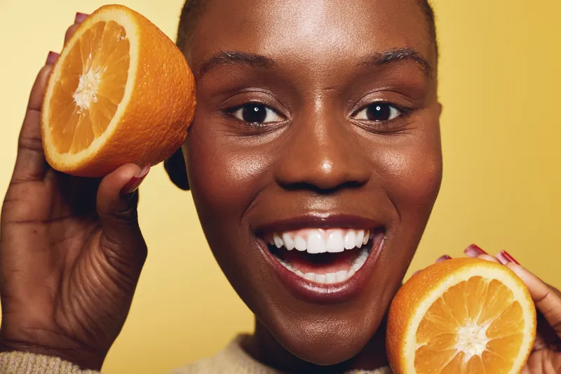 Close-up of happy African woman with dark skin holding two halves of fresh orange fruit near face. Bright yellow background and natural smile convey health, wellness, and freshness concept.