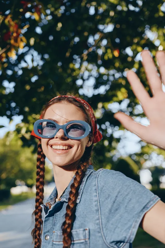 smiling woman with braids wearing blue sunglasses and denim shirt outdoors on sunny day with green trees and bright sunlight