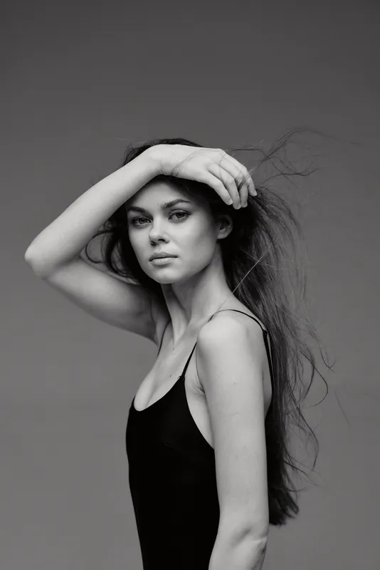 Black and white portrait of a young woman with long hair and a thoughtful expression wearing a black sleeveless top against a plain background.