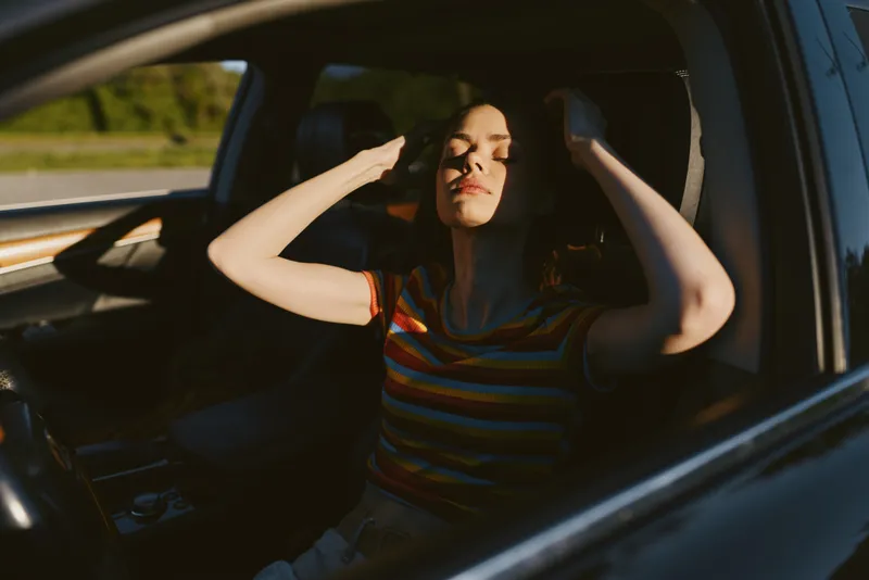 Woman wearing a striped shirt sits in a car, hands in her hair, sunlit interior, with warm, golden light. Close-up on soft retro film tones, conveying lifestyle, calm, and casual travel.