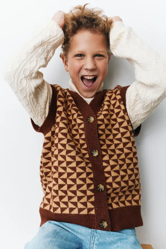 Energetic child poses joyfully with arms raised and a wide smile, wearing a geometric patterned vest over a cozy knit sweater, capturing playful confidence and happiness.