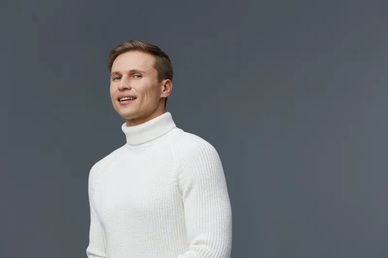 Confident young man with short brown hair wearing a white turtleneck sweater posing against a plain gray background studio portrait. People lifestyle concept.