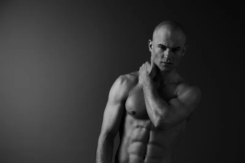 Black and white image of a muscular man posing thoughtfully, showcasing strength, emotion, and artistic expression in a minimalistic background.