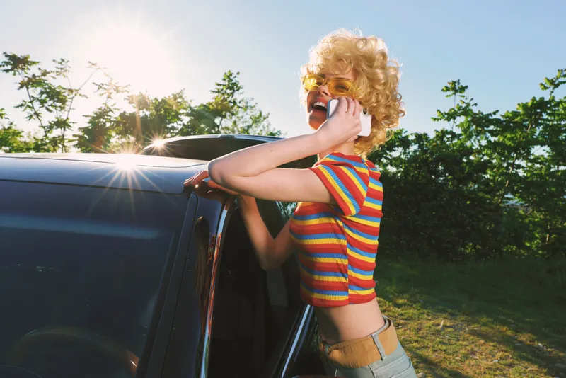 Casual woman leans against a vintage car, wearing a striped crop top and sunglasses, enjoying a sunny day with retro film color and a carefree outdoor lifestyle mood.