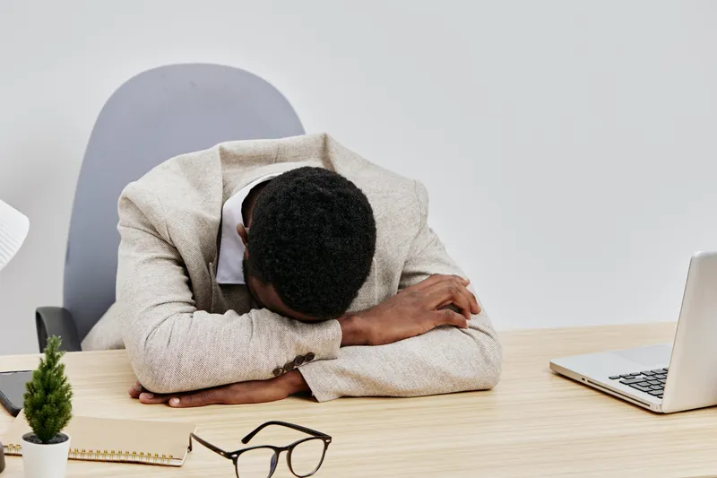 Frustrated man at desk lying face down on table in office environment, with laptop and glasses, showing stress or fatigue, concept of work pressure and exhaustion, minimalistic background