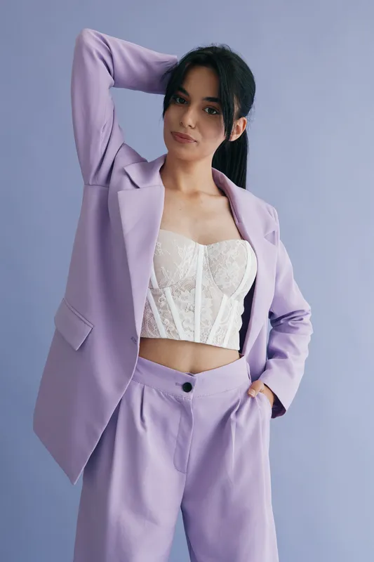 young woman in lilac suit and white corset top posing confidently against purple background with casual hairstyle and relaxed expression
