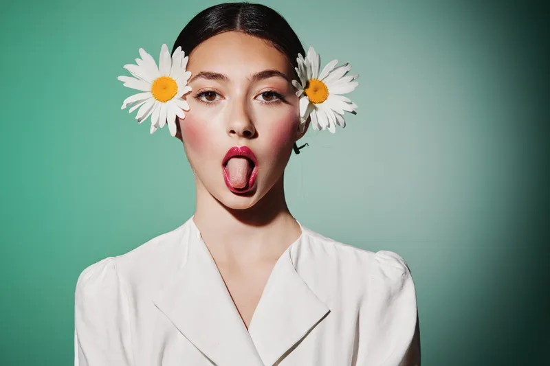 Young woman in white outfit with daisies in her hair sticks out tongue playfully on a teal background. Creative beauty and fashion concept with fresh and vibrant style.
