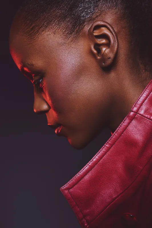 Profile of a young black woman with dark skin wearing a red leather jacket. The studio lighting highlights her smooth skin texture against a dark background. Concept of fashion and beauty.