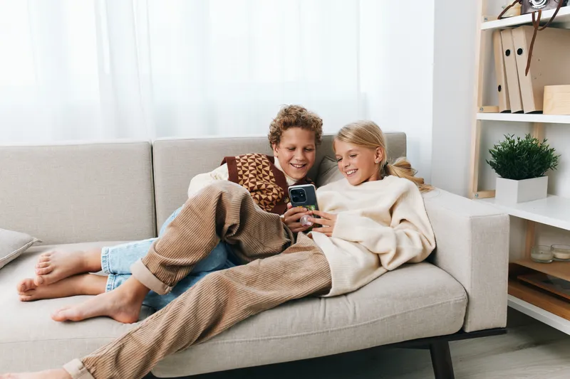 Two children lounge on a light sofa, smiling at a smartphone screen as they share a moment of playful connection and curiosity in a bright, cozy living room setting.