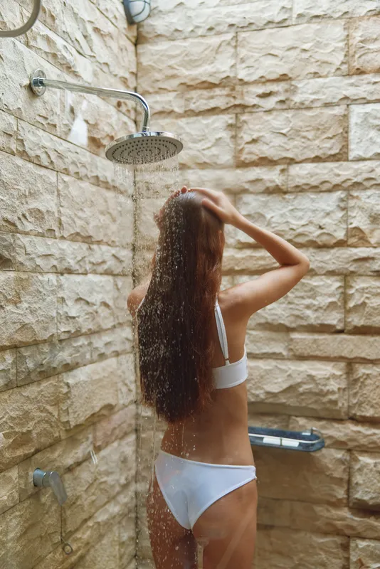 Woman showering outdoors under rainfall in a natural stone tiled shower area with relaxed posture and long wet hair, enjoying summer freshness and calm environment.