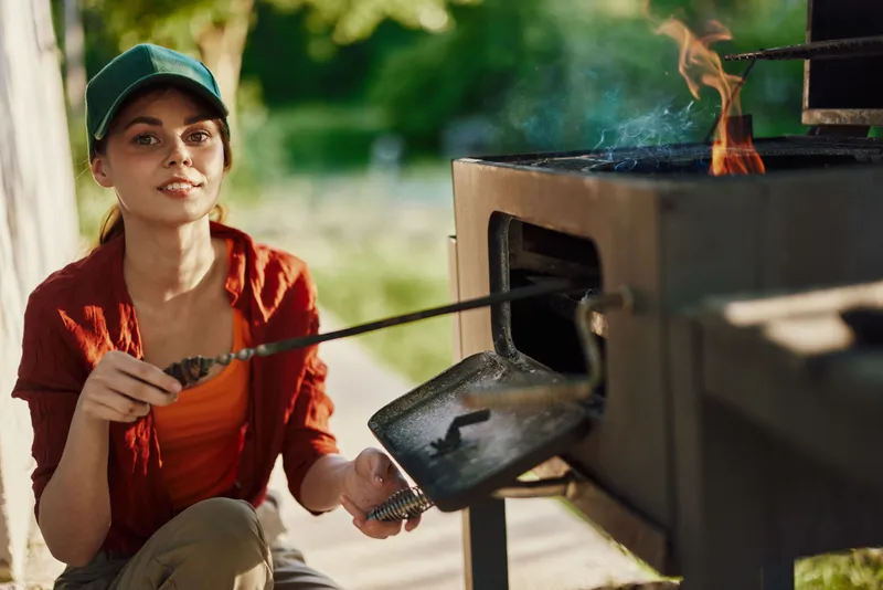 Young woman grilling outdoors with a cheerful expression, wearing casual summer clothes and a cap, surrounded by greenery in a sunny garden setting