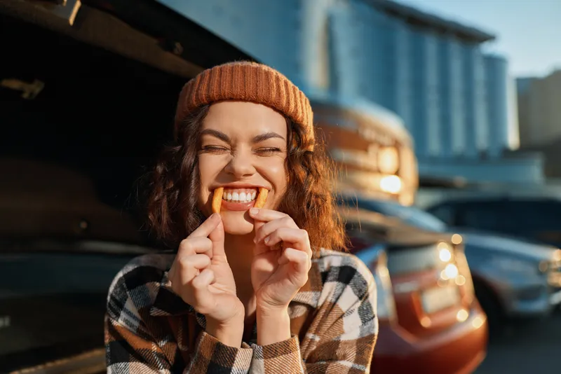 Woman holding fries like a playful smile, beanie on, leaning by a car in warm golden hour glow; candid authenticity and mindful living captured with intimate emotional storytelling.