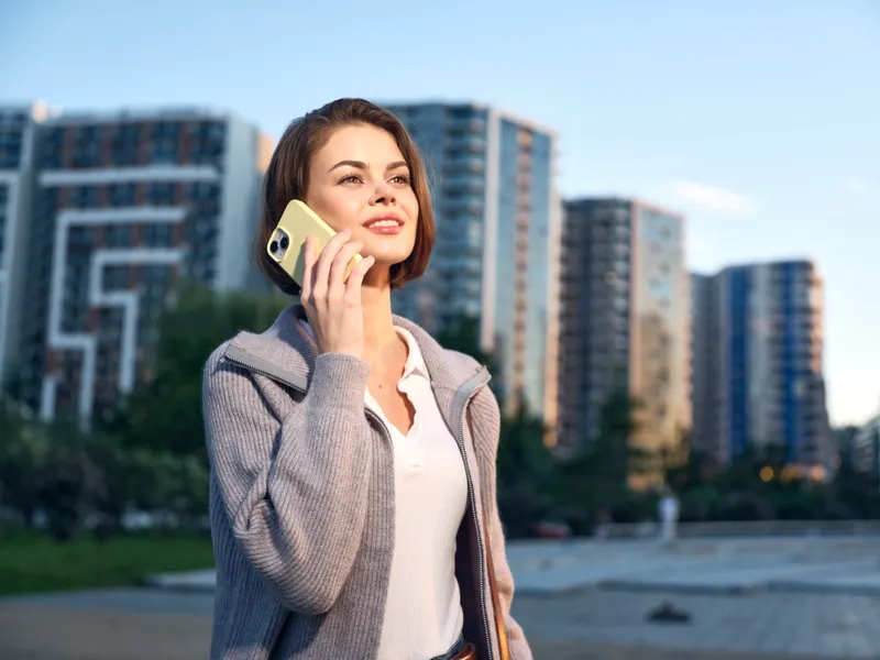 A confident business professional woman is seen outdoors on a smartphone in a modern urban setting. She wears business casual attire, smiles warmly, and exudes focus, energy, and a productive