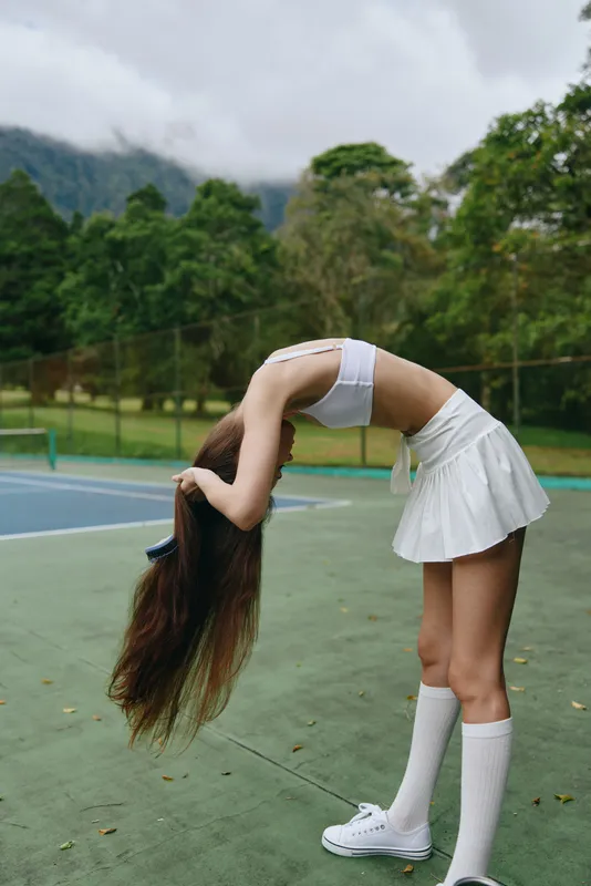 Young female tennis player adjusting her long hair on court, wearing a sporty white outfit that highlights her active lifestyle and vibrant energy in a lush green setting