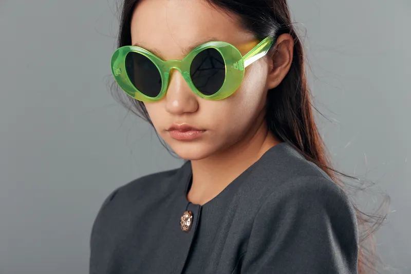 Woman with green sunglasses and long dark hair wearing dark gray clothing posing on gray background, stylish portrait with casual look and relaxed facial expression.