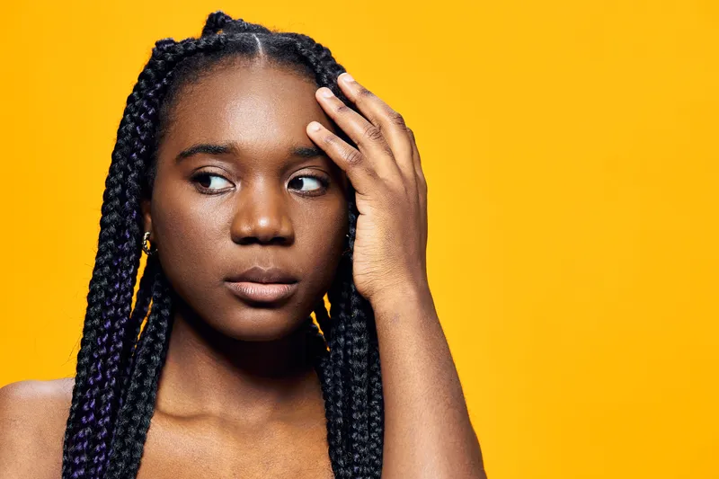 A young Black woman with braided hair looks thoughtfully away, showcasing a subtle expression against a vibrant yellow background in a bright and modern style