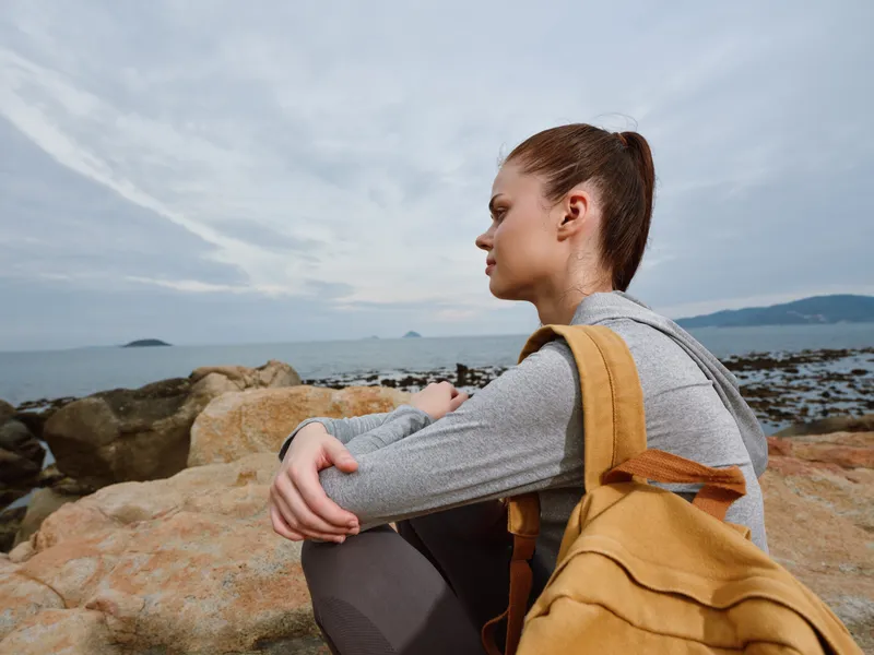 Young woman sitting on rocky beach, wearing gray long sleeve shirt and yellow backpack, contemplating serene ocean view, evoking tranquility and introspection