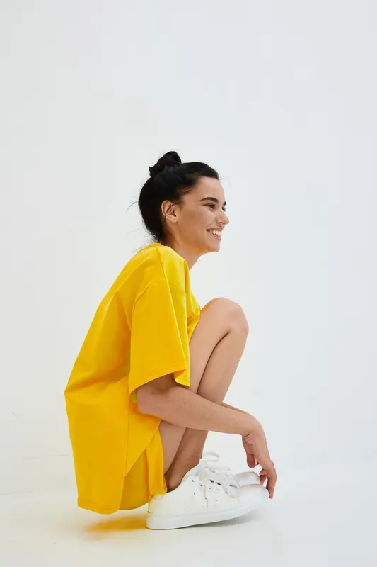 young woman in bright yellow oversized t-shirt sitting on floor smiling confidently with white sneakers on clean white background