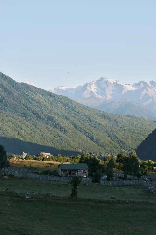 A tranquil rural landscape features rolling green hills, a wooden fence, a small house, and distant snow capped mountains under a clear blue sky.