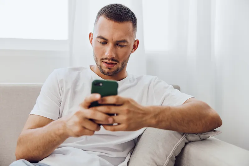 Focused man using smartphone, casually dressed, sitting on a sofa by the window, in a bright and modern interior, engaged with technology and lifestyle.