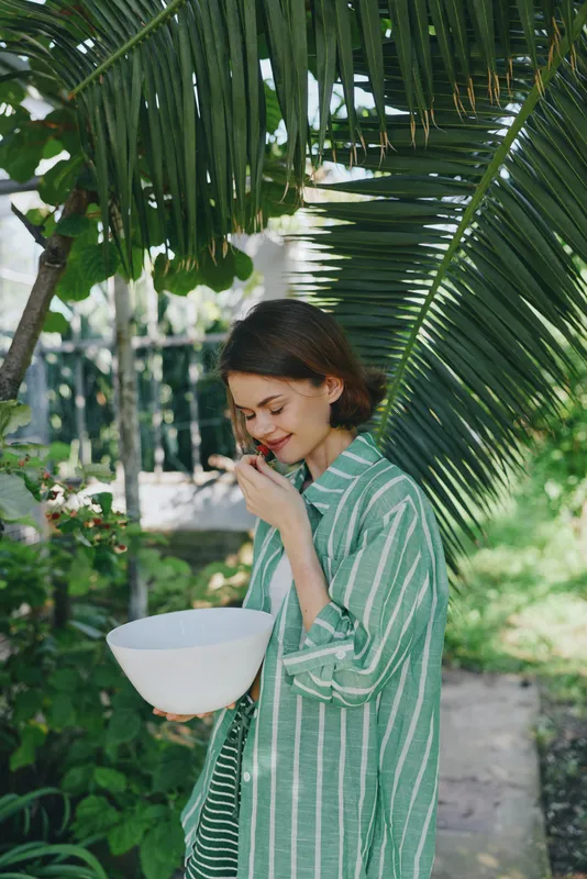 Young woman in a green striped shirt holds a white bowl under palm leaves, smiling as she tastes a snack.