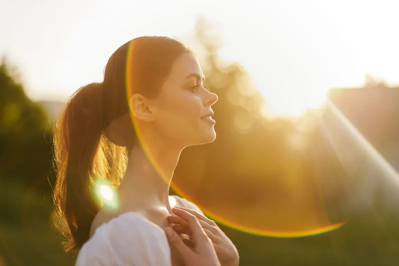 Young woman enjoying a sunny evening outdoors, radiating calm and positivity, with a soft ponytail and natural makeup, surrounded by a warm, glowing backdrop