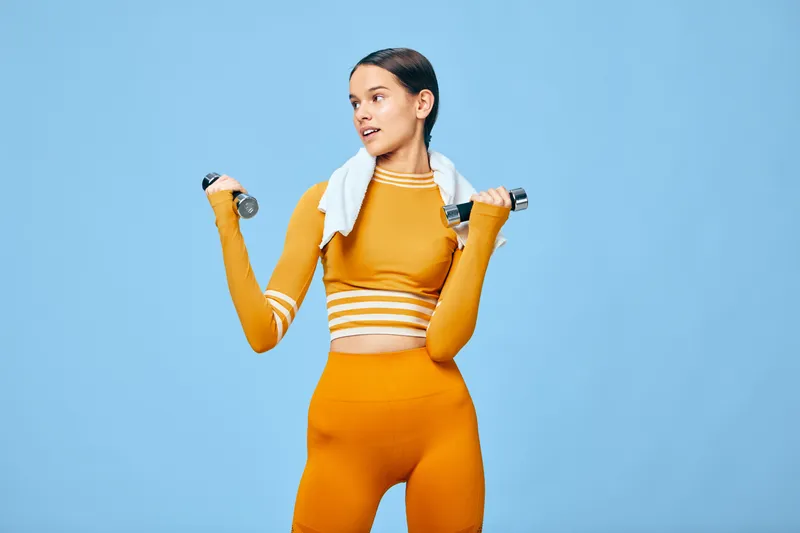 Athletic woman in bright orange sportwear lifts dumbbells with a towel over her shoulders against a blue backdrop, showcasing fitness, sport, workout, strength, balance and an active health lifestyle.