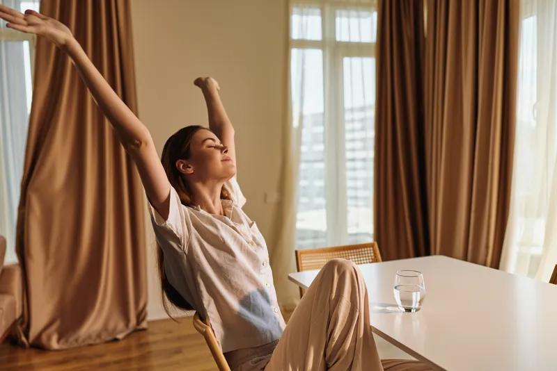 Woman stretching in morning at home, relaxation on chair by table with glass of water, sunlight through curtains, calm interior scene for wellness routine and peaceful comfort.