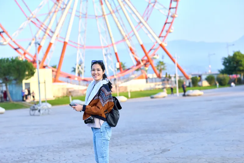 Woman traveler with backpack in an outdoor amusement park near a large ferris wheel, smiling while holding a tablet and scarf, casual fashion for leisure and exploration