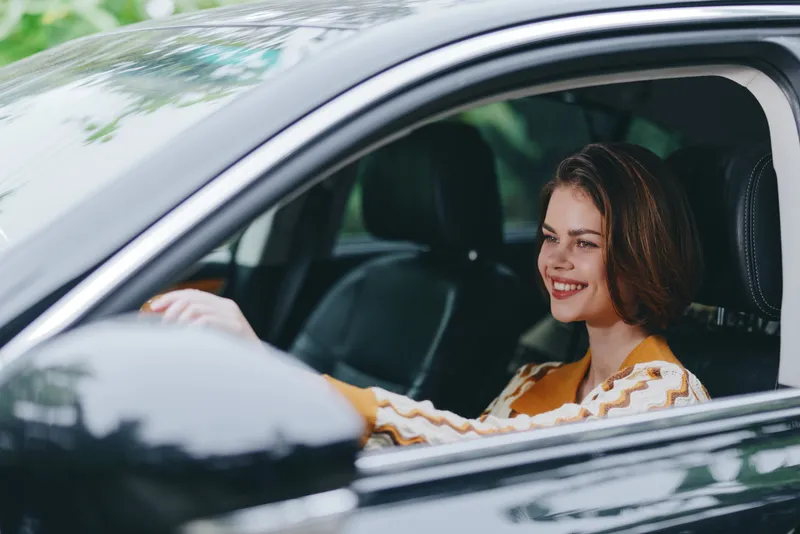 A smiling woman sits behind the wheel of a sleek car, enjoying a sunny drive and a relaxed moment in the interior.