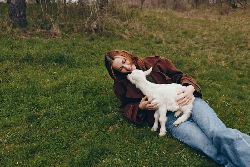 Young woman outdoors, sitting on grass, wearing a brown jacket and jeans, embracing a curious goat kid, tender moment, soft light, natural colors, tranquil countryside background, setting