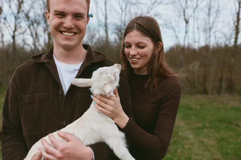 A joyful couple holds a playful goat kid in a sunlit field, smiling and calm. An affectionate moment outdoors in nature, capturing rural lifestyle and family bonding.