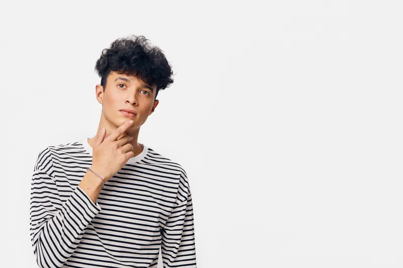 thoughtful man wearing striped shirt looks curious and reflective against a plain white background, young adult with curly hair expressing contemplation and focus.