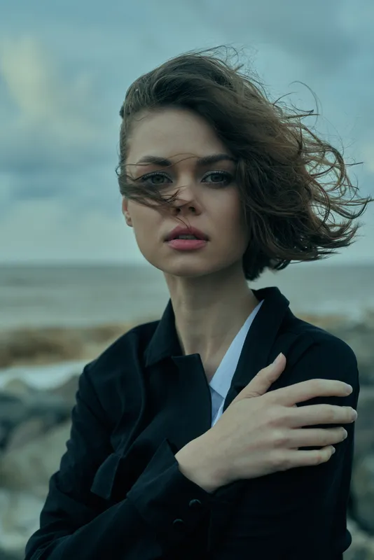 Woman on a windy beach wearing a dark blazer, hair blown by sea breeze, confident pose and moody coastal fashion for outdoor storytelling