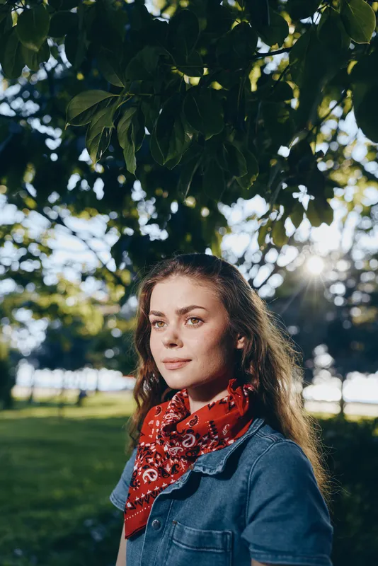 lifestyle woman in modern boho-western denim street style wearing red bandana outdoors with natural light and film color tones, calm expression under green leaves
