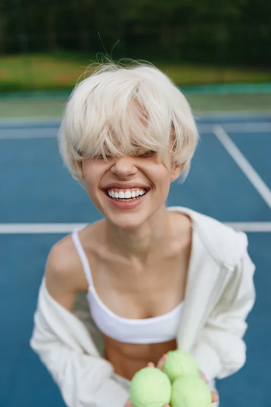 Cheerful young woman with short blonde hair in sportswear on a tennis court holding tennis balls, radiating joy and energy in a vibrant outdoor setting