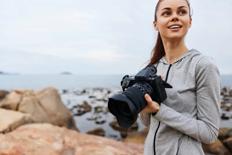 Young woman holding a camera by the shoreline, dressed in casual gray sportswear, exuding a cheerful expression amidst a scenic coastal background