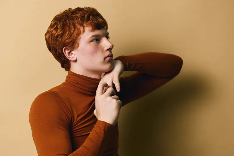 Young man with red hair wearing brown turtleneck adjusting sleeve against neutral background. Portrait of confident male model in casual clothing with thoughtful expression and side view.