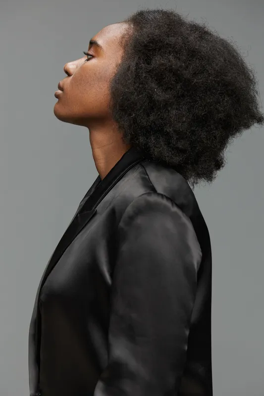 Profile portrait of a Black woman with textured hair in a black blazer, showcasing confidence and elegance against a muted gray background