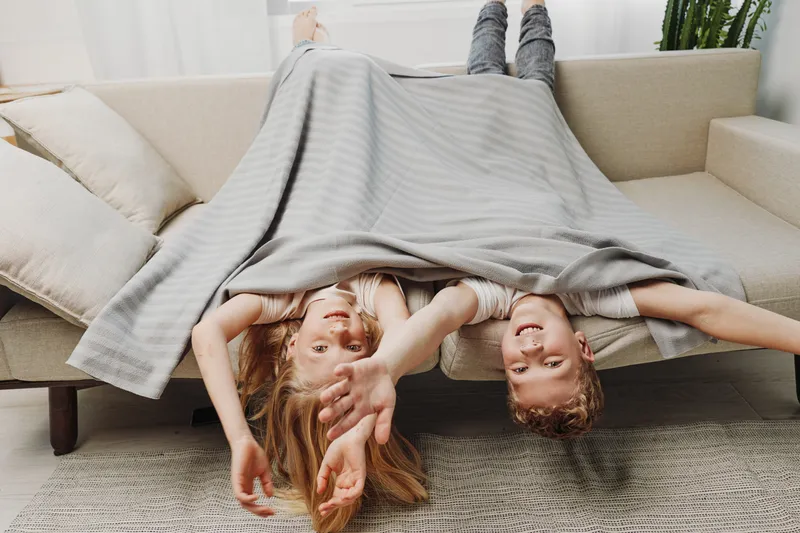 Two happy children lying upside down on the sofa covered with a blanket, smiling and playing indoors in a cozy living room environment with natural light.