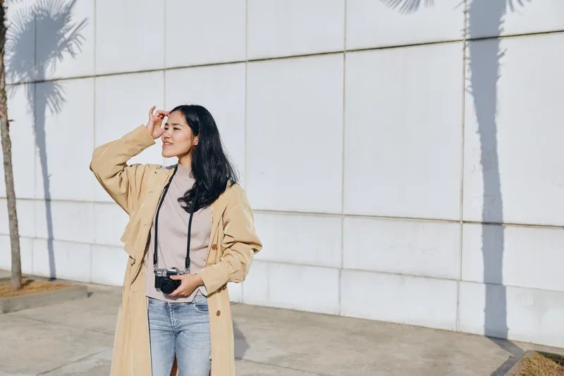 Woman with camera in hand, outdoors fashion shoot featuring a trench coat, street style and sunlight, capturing urban lifestyle and creative photography, art.