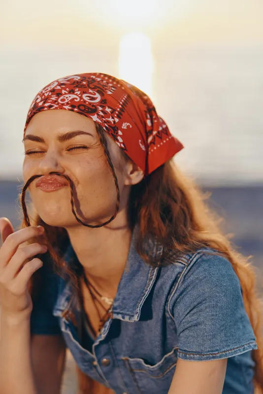 lifestyle woman in modern boho-western denim street style, playful expression, red bandana, warm film color tones, casual outdoor fun near water at sunset