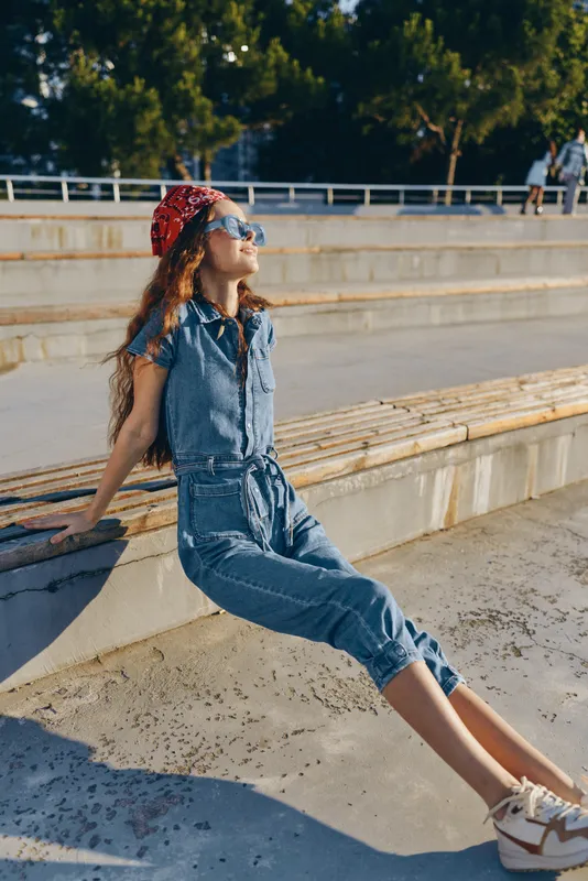 Lifestyle woman in modern boho-western denim street style sits relaxed under warm sunlight, wearing sunglasses and a red bandana, film color tones enhance the mood.
