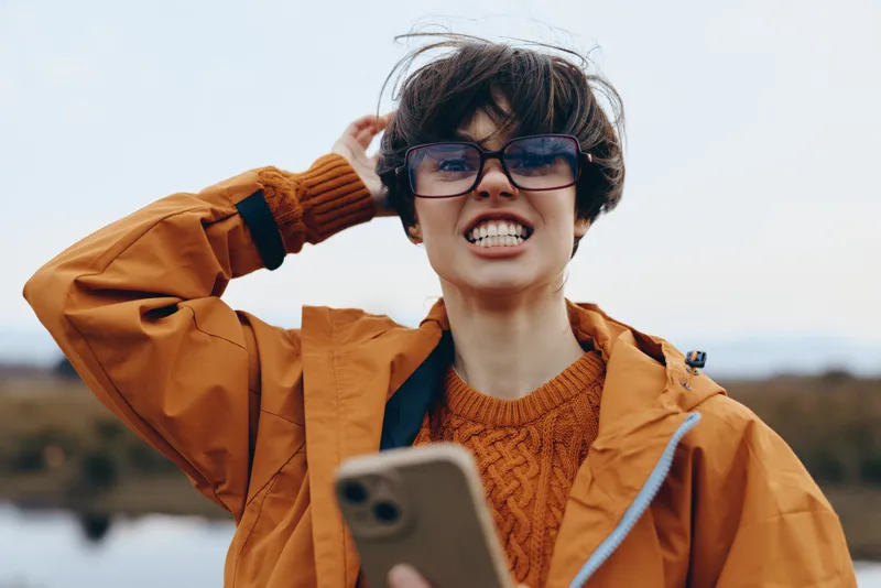 Woman outdoors smiling, wearing glasses, orange jacket and knit sweater, holding phone selfie, autumn daylight, wind in hair, portrait lifestyle, blurred landscape background