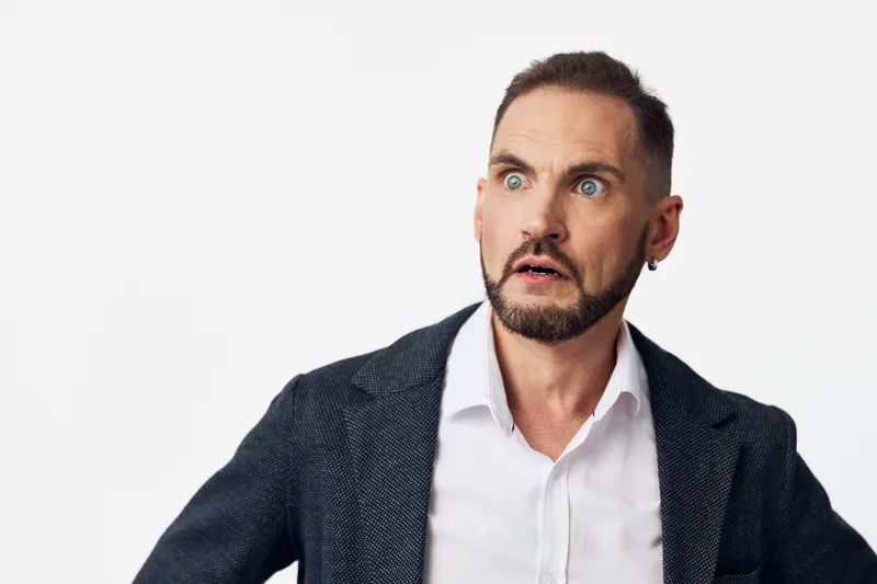 A business professional man stands against a solid color background, displaying a surprised look that conveys curiosity, alertness, and dynamic momentum in a corporate scene.